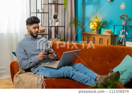 Excited Indian man playing video game on laptop computer spending leisure time at home couch Excited Indian man playing video game on laptop computer spending leisure time at home couch 112356874