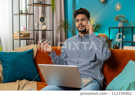 Hispanic young man making phone conversation call with client or colleague sitting at home couch Hispanic young man making phone conversation call with client or colleague sitting at home couch 112356902