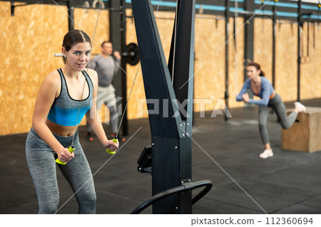Slender athletic girl work out muscles of arms and shoulders on rope loop simulator in gym of fitness club 112360694