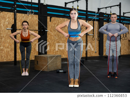 Strong woman using resistance band in exercise routine in crossfit gym. Group of people performs fitness exercises with stretch rope on background Strong woman using resistance band in exercise routine in crossfit gym. Group of people performs fitness exercises with stretch rope on background 112360853