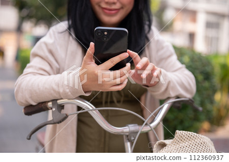 A beautiful, happy young Asian woman in a cute dress is using her smartphone on her bike in the city 112360937