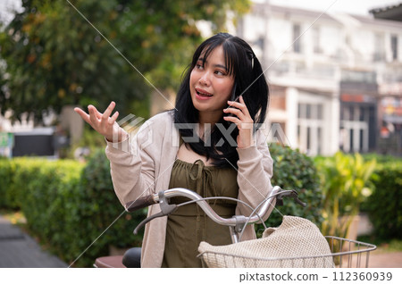 A beautiful, happy young Asian woman is talking on the phone while riding a bike in the city. 112360939