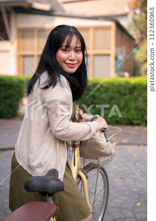 A smiling, happy young Asian woman in a cute dress is riding a bicycle in the city 112360968