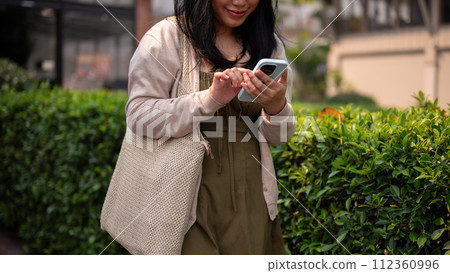 A happy Asian woman in casual clothes using her smartphone while walking in the city on a bright day A happy Asian woman in casual clothes using her smartphone while walking in the city on a bright day 112360996
