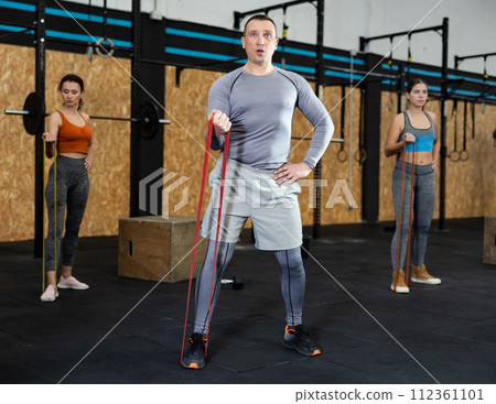 Strong man using resistance band in exercise routine in crossfit gym. Group of people performs fitness exercises with stretch rope on background 112361101