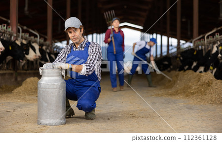 Male farm worker carrying big milk can walking in cowshed on dairy farm 112361128