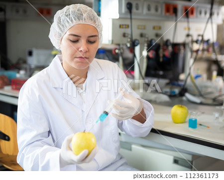 Young female scientist checking fruits for harmful elements, injecting syringe into apple in lab 112361173