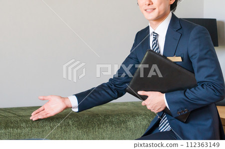 一位來自房屋建築商的年輕男性推銷員穿著西裝,在樣品屋的客廳展示並推薦沙發。 一位來自房屋建築商的年輕男性推銷員穿著西裝,在樣品屋的客廳展示並推薦沙發。 112363149