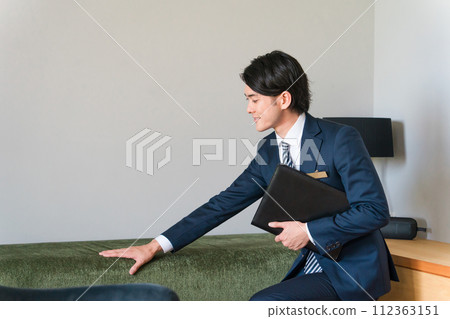 A young male salesman from a house builder wearing a suit shows and proposes sofas in the living room of a model house. A young male salesman from a house builder wearing a suit shows and proposes sofas in the living room of a model house. 112363151
