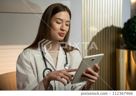 Female Doctor in White Coat Reviewing Patient Information on a Tablet in Her Office During the Day Female Doctor in White Coat Reviewing Patient Information on a Tablet in Her Office During the Day 112367466