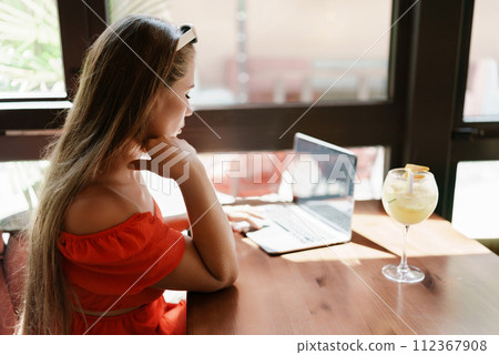 Woman works on laptop in a sunny cafe, glass of juice nearby. The setting is modern, with natural light illuminating the space. The image captures a moment of focused work. 112367908