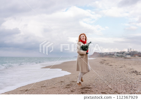 Redhead woman Christmas tree sea. Christmas portrait of a happy redhead woman walking along the beach and holding a Christmas tree in her hands. Dressed in a light coat, white suit and red mittens. Redhead woman Christmas tree sea. Christmas portrait of a happy redhead woman walking along the beach and holding a Christmas tree in her hands. Dressed in a light coat, white suit and red mittens. 112367929
