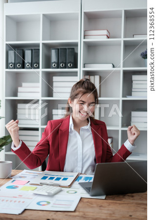 Beautiful asian businesswoman working on laptop and calculating with financial calculator from statistics data graphs, charts. Successful business results in modern office wear red shirt. 112368554