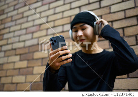 Smiling young Asian man using mobile phone and listening to music with headphones against a wall Smiling young Asian man using mobile phone and listening to music with headphones against a wall 112371405