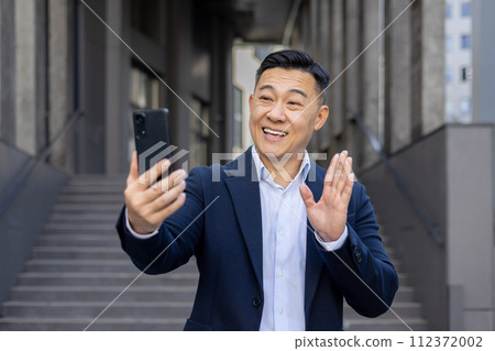 Cheerful Asian businessman engaging in a video call using his smartphone, smiling and waving hello outdoors. 112372002