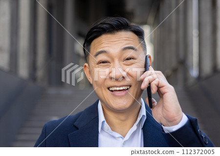 Cheerful Asian businessman casually dressed in a suit making a happy phone call while standing outdoors. 112372005