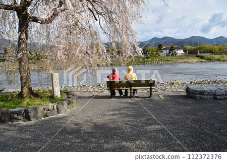 日本春天的京都,嵐山週邊的街道,旅遊勝地桂川,櫻花盛開。一對老夫婦坐在長椅上賞櫻花。 日本春天的京都,嵐山週邊的街道,旅遊勝地桂川,櫻花盛開。一對老夫婦坐在長椅上賞櫻花。 112372376