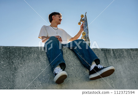 Low angle of young male skateboarder holding skateboard sitting on ramp in skate park Low angle of young male skateboarder holding skateboard sitting on ramp in skate park 112372594