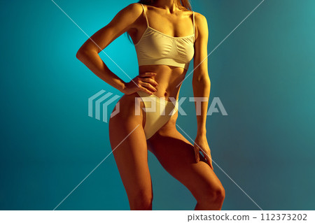 Cropped portrait of young slim woman in white underwear posing holds hands gently on leg and hip in neon light against studio background. 112373202