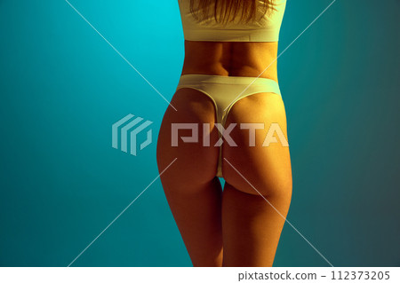 Rear view of female model in underwear showing back, buttocks without cellulite in yellow neon light against gradient studio background. 112373205
