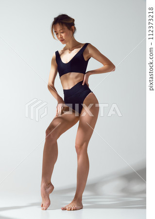 Confident, beautiful, slim woman in black swimwear posed thoughtfully against grey studio background in sun light. Protect skin from sun. 112373218