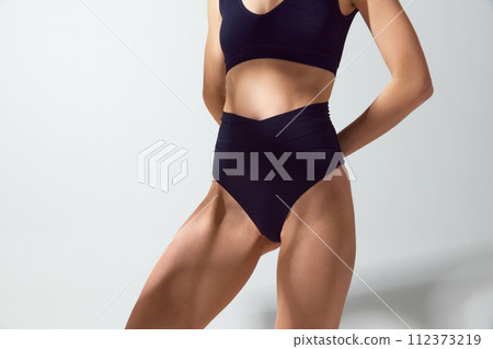 Close up of young woman in blue swimwear holds hands behind back against grey studio background in sun light. Protect skin from sun. 112373219