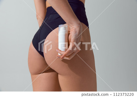 Close-up of woman's hand holding a skincare product near her thigh against grey studio background. Body care and beauty routines. 112373220