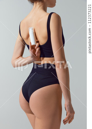 Rear view portrait of young fit woman in blue underwear holds jar of cosmetic product behind back against grey studio background. 112373221