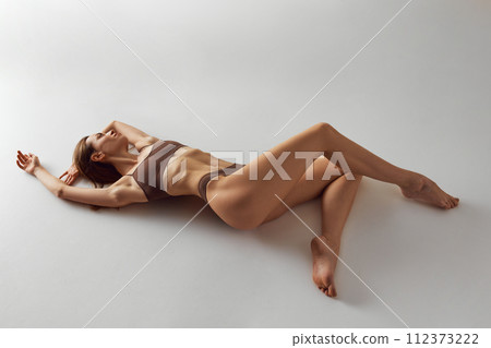 Beautiful, slim woman in relaxed pose on floor, stretching in her brown underwear against grey studio background. 112373222