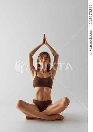 Mindfulness. Focused, calm woman in brown underwear sitting in lotus pose and raising hands with closed eyes against grey studio background. 112373232