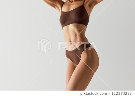 Cropped portrait of female model posing against grey studio background. Toned midriff of woman in chocolate brown underwear set. 112373252