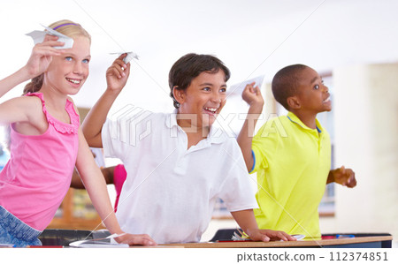 Happy kids, students and paper plane in classroom on break at desk for fun games to throw or relax. Diversity, distraction or children playing with smile, origami jet or airplane in school together 112374851