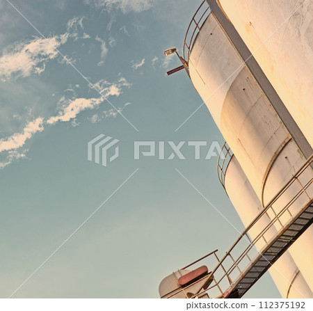 Closeup, silo and tank at plant for agriculture, bulk storage and product safety with sky, clouds and outdoor. Container, agricultural structure and oil refinery tower with low angle for distillery 112375192