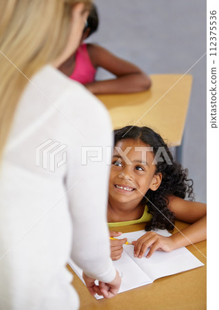 Teacher woman, classroom and help girl at desk with notebook, advice and knowledge for education. People, learning and development with person, child and check book for progress, school or academy 112375536
