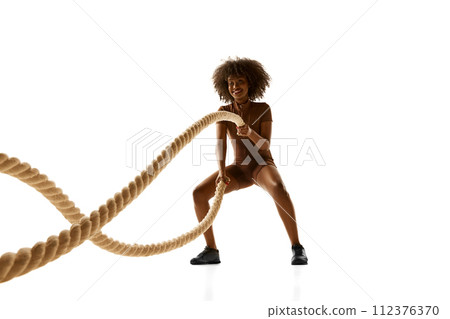 Smiling African-American woman in brown sports suit using thick ropes for fitness training against white studio background. 112376370