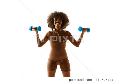 Smiling African-American woman lifting blue dumbbells, dressed in brown workout suit against white studio background. 112376445