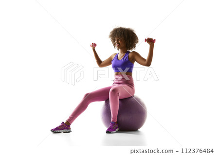 African-American woman in purple sportswear sitting on exercise ball, lifting small weights against white studio background. 112376484