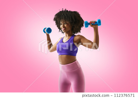 Smiling woman with dumbbells raised, wearing a purple sports bra and leggings against gradient pink studio background. Smiling woman with dumbbells raised, wearing a purple sports bra and leggings against gradient pink studio background. 112376571