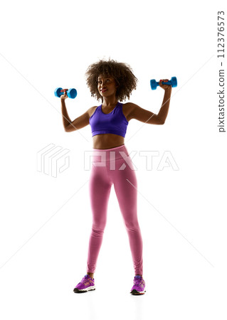 Smiling woman with dumbbells raised, wearing a purple sports bra and leggings against white studio background. Smiling woman with dumbbells raised, wearing a purple sports bra and leggings against white studio background. 112376573