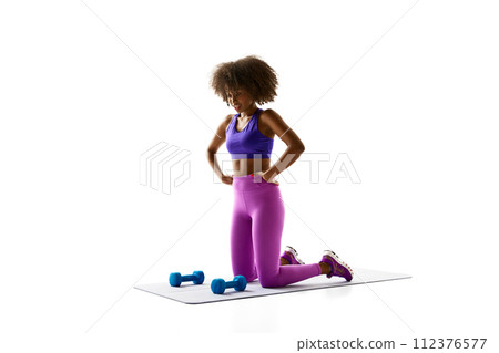 Woman in purple activewear kneeling on mat beside blue dumbbells, looking down against white studio background. Woman in purple activewear kneeling on mat beside blue dumbbells, looking down against white studio background. 112376577