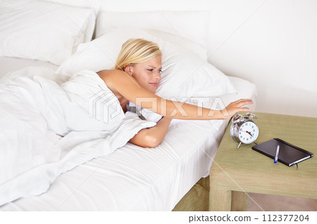 Snooze, alarm and woman wake up with clock in morning and tired on bed in home with stress, fatigue and lazy. Angry, girl and press stop or time to start day and frustrated with routine in bedroom 112377204