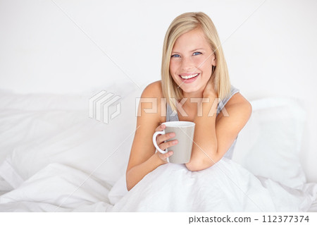 Woman, portrait and drinking coffee in bed for morning relax or home beverage for comfort, peace or happy. Female person, face and smile with mug on duvet for warm caffeine or vacation, rest or tea 112377374