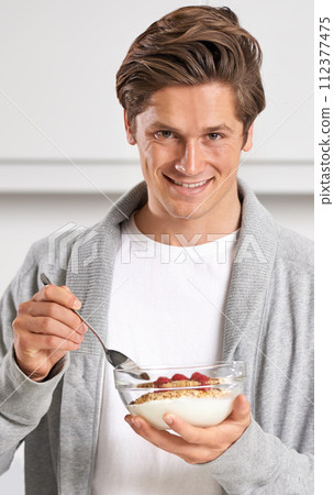 Smile, portrait and man with cereal in kitchen of home for hydration, health and wellness. Happy, confident and young male person eating breakfast with fruit, yoghurt and muesli in modern apartment. 112377475