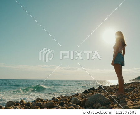 A woman stands on a rocky beach, looking out at the ocean A woman stands on a rocky beach, looking out at the ocean 112378595