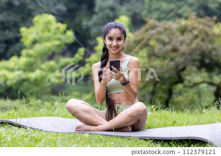 Young Indian woman in a tracksuit uses her phone while sitting on a yoga mat in a serene park, embodying fitness and relaxation. 112379121