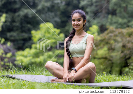 Young Indian woman in a tracksuit enjoys a calm yoga session in a green park, embodying peace and healthy lifestyle. Young Indian woman in a tracksuit enjoys a calm yoga session in a green park, embodying peace and healthy lifestyle. 112379132