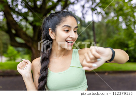 Motivated Indian woman in a tracksuit uses a fitness tracker while running in a lush green park, embodying health and vitality. Motivated Indian woman in a tracksuit uses a fitness tracker while running in a lush green park, embodying health and vitality. 112379155