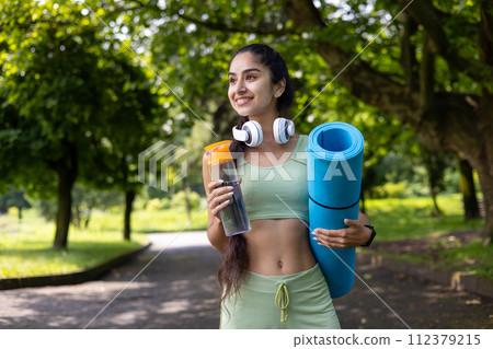 A fitness-focused Indian woman in workout attire holds a yoga mat and water bottle while enjoying a park. 112379215