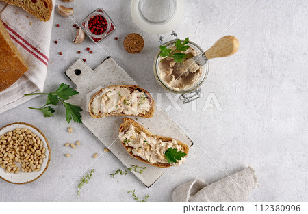 Homemade pate, spread or mousse in glass jar with sliced bread and herbs with pine nuts, light concrete background. Homemade pate, spread or mousse in glass jar with sliced bread and herbs with pine nuts, light concrete background. 112380996