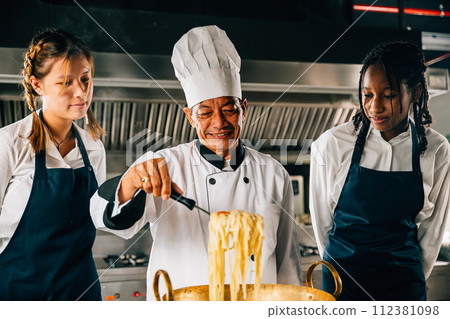 Chef guides students in kitchen. Schoolgirls cook Japanese noodle. Kids with teacher at stove. Smiling portrait of learning is modern education. Making dinner with ladle gives joy. 112381098